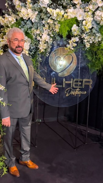 Homem em traje social ao lado de uma placa com o nome 'ILHER D'Osseus', decorada com flores.