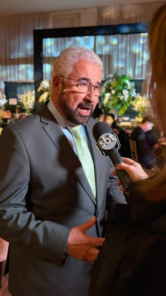 Homem de terno cinza com gravata verde, sendo entrevistado em um evento, segurando um microfone.