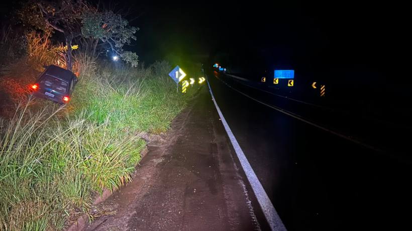 Carro acidentado à beira da estrada escura, com sinais de curva visíveis e vegetação ao redor.