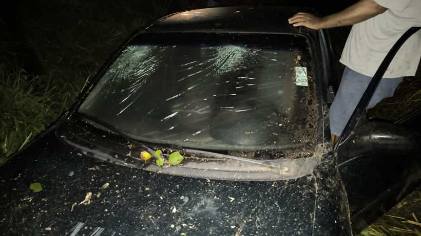 Um carro preto coberto de poeira e detritos, estacionado em um local afastado à noite, com uma pessoa ao lado.