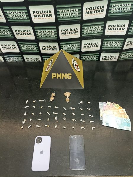 Mesa com objetos relacionados a uma apreensão policial, incluindo drogas em embalagens, dinheiro em notas variadas, e dois celulares, em frente a um fundo com placas da Polícia Militar.