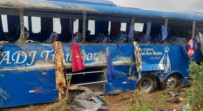 Ônibus danificado com janelas quebradas, coberto de sujeira, e roupas penduradas nas laterais. O veículo é azul e apresenta inscrições na lateral.