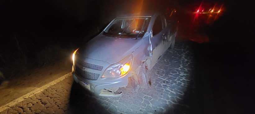 Um carro acidentado à noite, com danos visíveis na lateral e faróis acesos, estacionado em uma estrada de paralelepípedos.