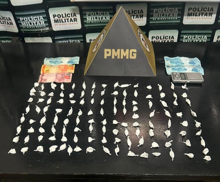 Um tablete em forma de pirâmide com a inscrição 'PMMG', ao lado de diversas porções brancas dispostas em fileiras, notas de dinheiro em reais e uma balança de precisão sobre uma mesa, com fundo de parede da polícia militar.