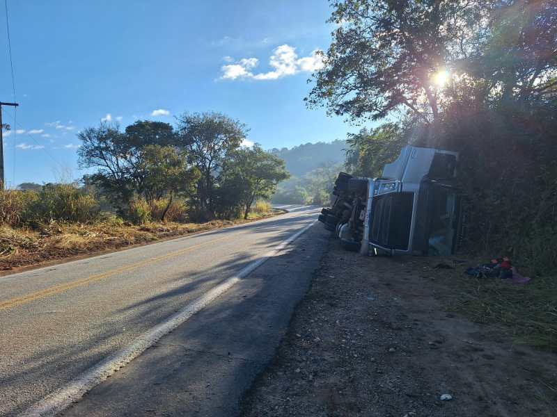 Caminhão tombado ao lado da estrada, cercado por vegetação e sob um céu claro.