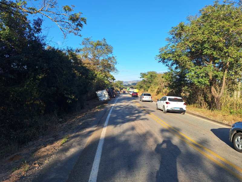 Caminho rural com carros passando ao lado de um caminhão tombado, cercado por vegetação.