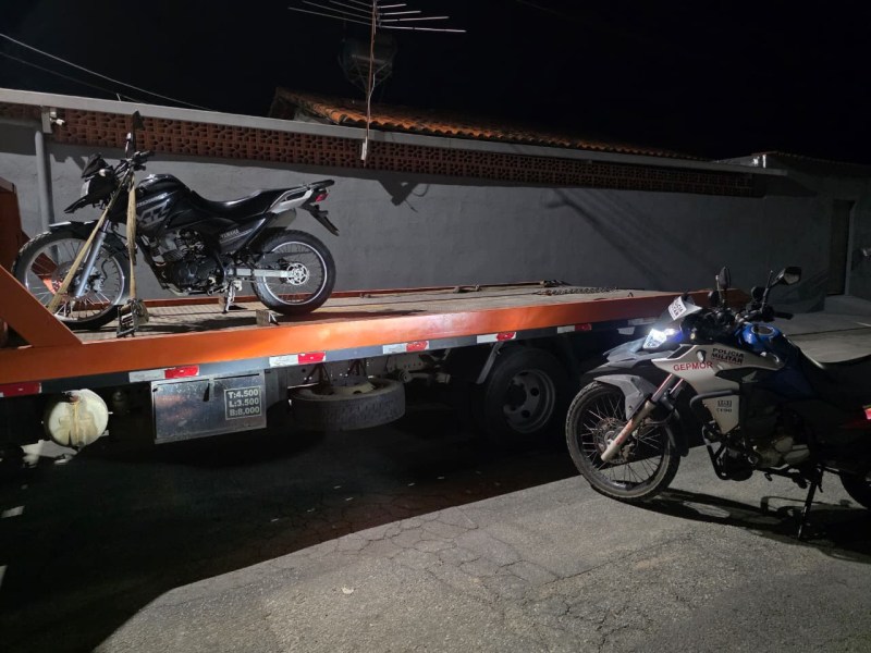 Uma moto sendo carregada em um caminhão guincho à noite, com outra moto estacionada próxima.