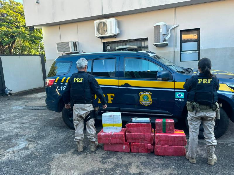 Dois agentes da PRF em frente a uma viatura, ao lado de pacotes suspeitos empilhados no chão.
