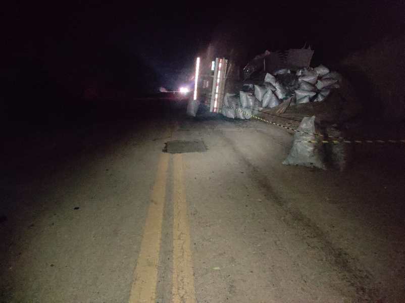 Caminho bloqueado por caminhão tombado, com sacos empilhados e sinalização de segurança na estrada à noite.