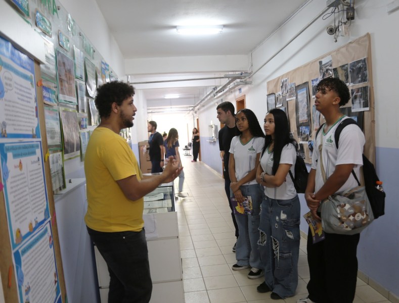Um educador falando com um grupo de estudantes em um corredor escolar, com painéis informativos nas paredes.