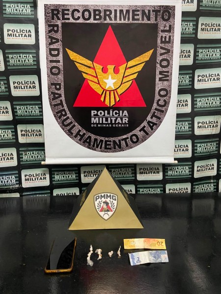 Foto de uma mesa com diversos itens, incluindo um cartaz da Polícia Militar, uma pirâmide com o logo da PM, dois bills de dinheiro e substâncias não identificadas em um pano.