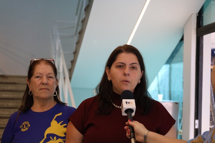 Duas mulheres em um ambiente interno, uma delas sendo entrevistada com um microfone em mãos, enquanto a outra ouve atentamente ao fundo.