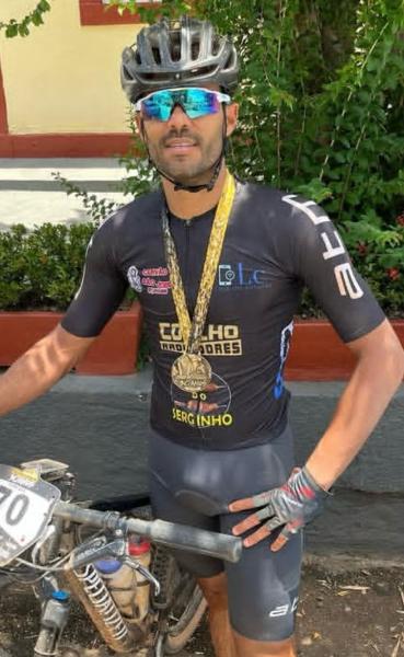 Ciclista posando com medalha de ouro e bicicleta, vestindo roupa de competição e óculos de sol.