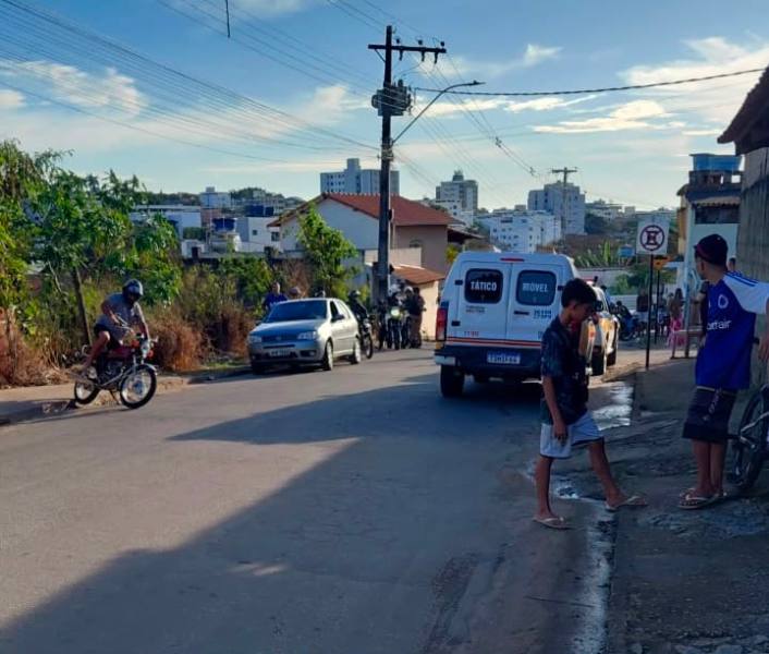 Rua com vegetação, carros estacionados e duas crianças interagindo. Um ciclista passa ao fundo. Edifícios visíveis ao longe.