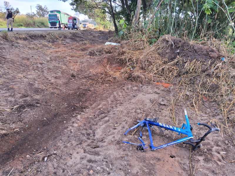 Quadro de bicicleta azul abandonado em terreno de terra ao lado da estrada, com veículos ao fundo.