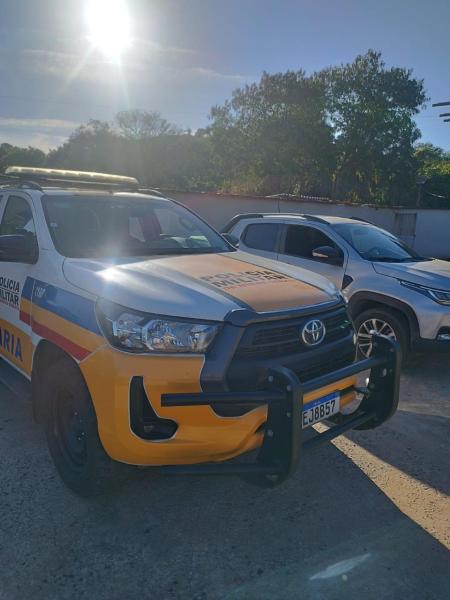 Veículo da polícia com pintura amarela e azul estacionado, ao lado de um carro de passeio, sob um céu claro e ensolarado.