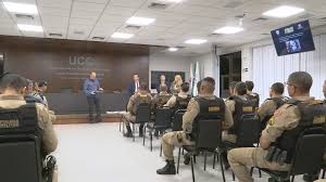 Reunião de policiais em uma sala, com alguns deles sentados e outros em pé apresentando informações.