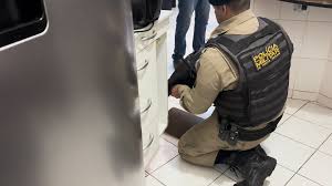 Policial agachado em uma cozinha, examinando algo próximo ao fogão enquanto uma pessoa está de pé ao lado.