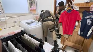 Dois homens em trajes de serviço verificando uma máquina de lavar em um ambiente doméstico, com roupas penduradas e uma bancada ao fundo.