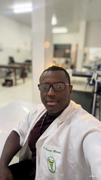 A man wearing a white lab coat with a stethoscope around his neck, smiling at the camera in a medical office setting.