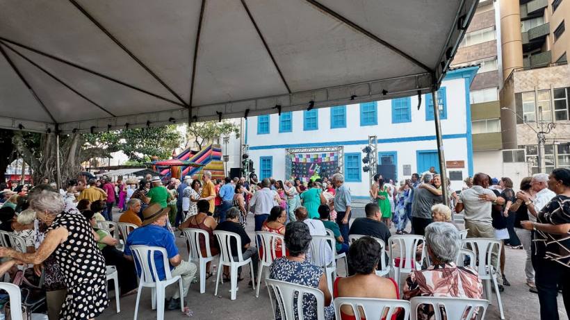 Evento ao ar livre com um grande grupo de pessoas dançando sob uma tenda, enquanto outros assistem. Ao fundo, um edifício azul e uma decoração festiva.