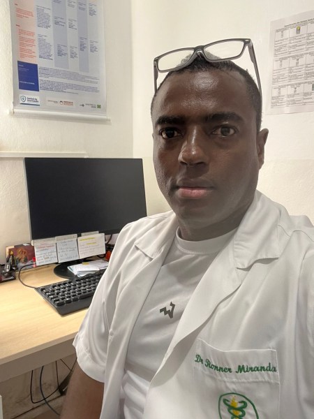 A healthcare professional wearing a white lab coat is sitting in an office with a computer desk in the background. The person has glasses resting on their head and appears focused.
