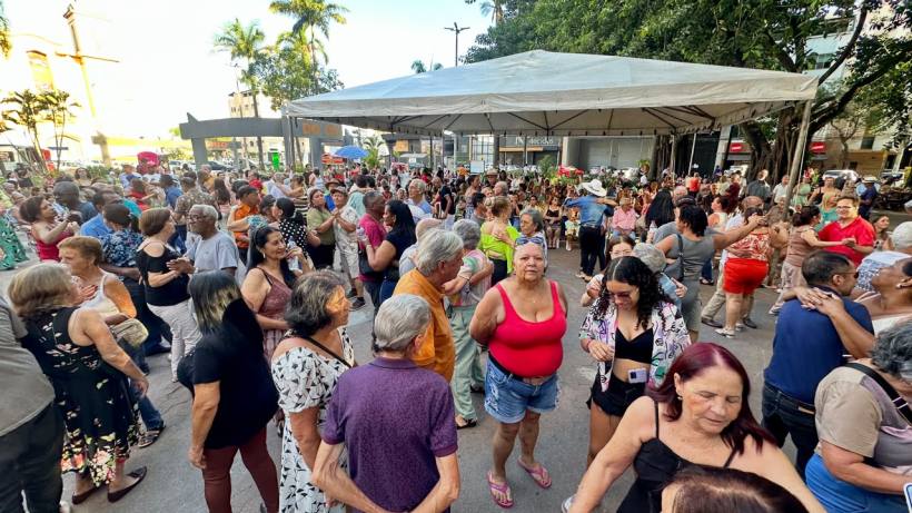 Grupo de pessoas idosas dançando em um evento ao ar livre sob um toldo, com árvores e edifícios ao fundo.