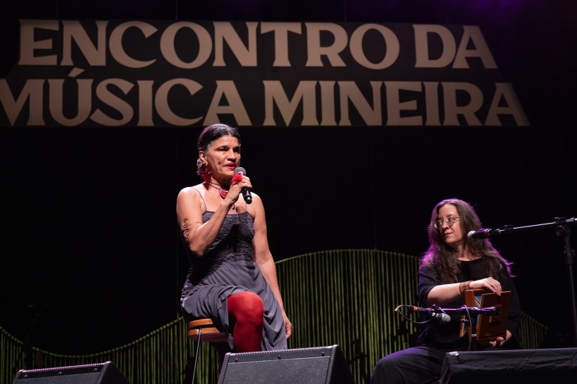 Artista se apresentando em palco durante o Encontro da Música Mineira, ao lado de uma musicista com um instrumento.