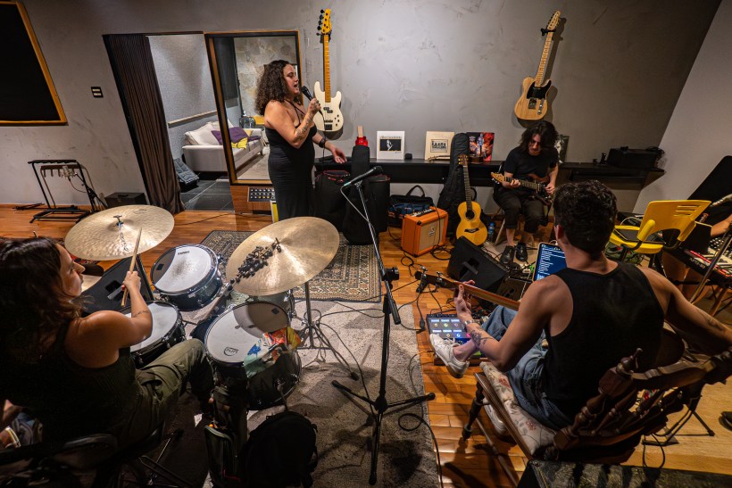 Banda ensaiando em um estúdio, com uma vocalista de pé, um baterista à esquerda e dois guitarristas sentados. Instrumentos musicais e equipamentos estão espalhados pelo ambiente.