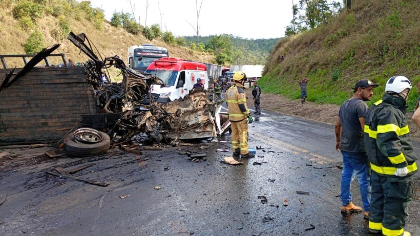 Acidente de trânsito na estrada com caminhão destruído, equipe de bombeiros, ambulância e pessoas ao redor.