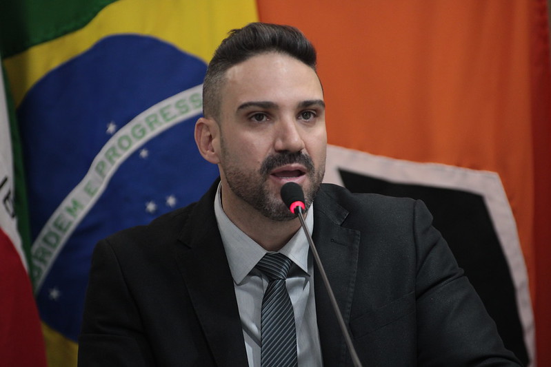 Homem falando em um microfone, com expressão séria, diante de bandeiras do Brasil e de outros países ao fundo.