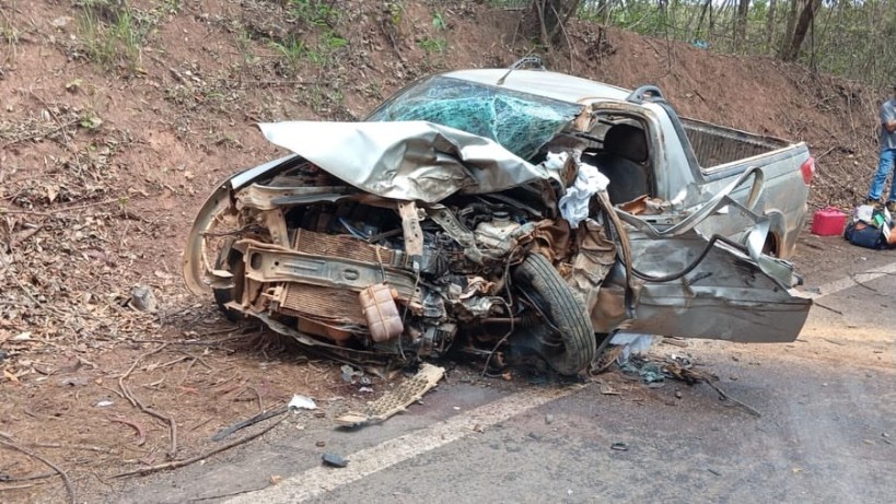 Imagem de um carro acidentado, com a frente esmagada e danos significativos, encostado em uma encosta ao lado da estrada.