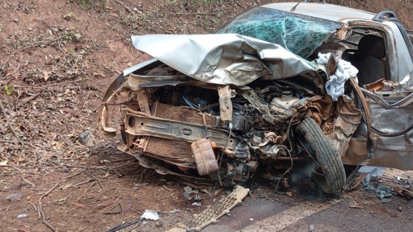 Carro acidentado com a parte da frente severamente danificada, localizado ao lado da estrada em um terreno de terra.