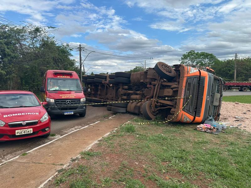 Caminhão tombado na estrada, com veículos dos bombeiros ao lado e sinalização de segurança.