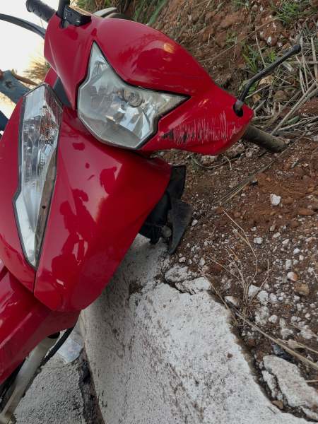 Uma motocicleta vermelha em ângulo, com evidências de danos na parte frontal e no farol, estacionada próximo a um bordão de concreto.