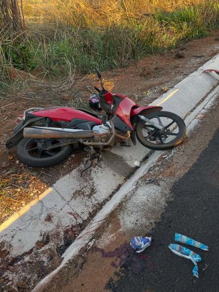 Motocicleta caída em uma calçada, ao lado de uma pista de asfalto, com sujeira ao redor e um par de roupas perto.