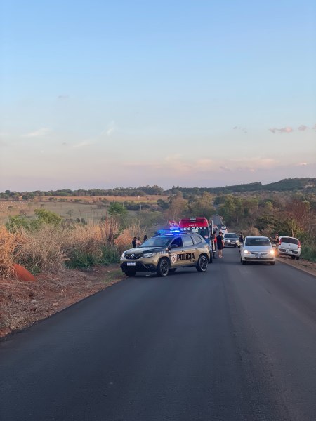 Veículo da polícia com sirenes acionadas em uma estrada rural, com outros carros parados ao fundo e um cenário de vegetação.