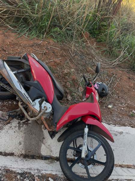 Uma motocicleta vermelha caída ao lado de uma estrada, com grama alta ao fundo.