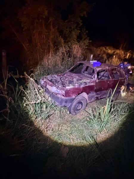 Um carro enferrujado e danificado estacionado em uma área coberta de grama alta, iluminado por uma lanterna durante a noite.
