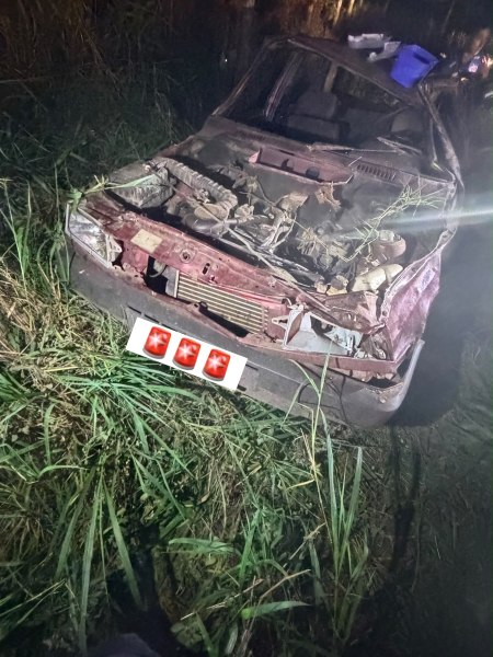 Um carro abandonado e danificado em meio à grama alta, com o capô aberto e peças visíveis.