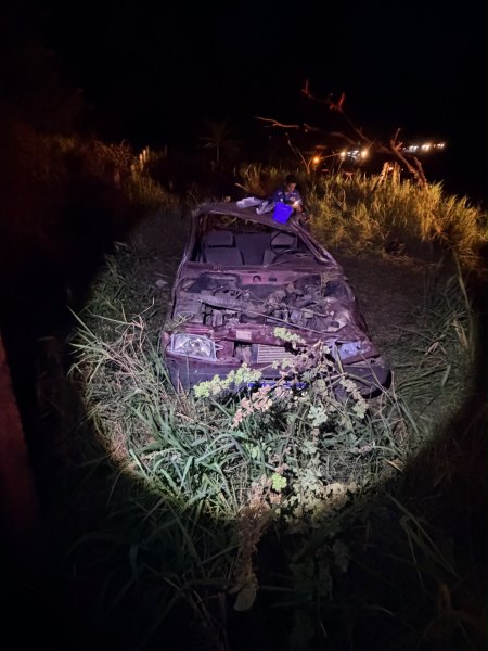 Um carro danificado abandonado em um campo com grama alta à noite, iluminado por uma lanterna.