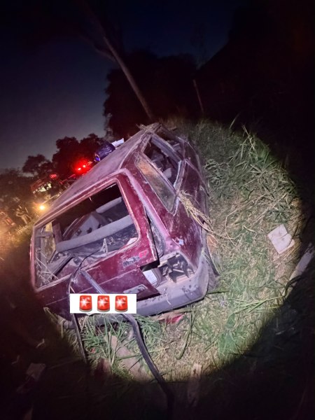 Carro acidentado em um campo à noite, coberto de grama e detritos.