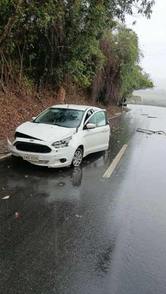 Um carro branco acidentado na beira da estrada em um dia chuvoso, cercado por vegetação.