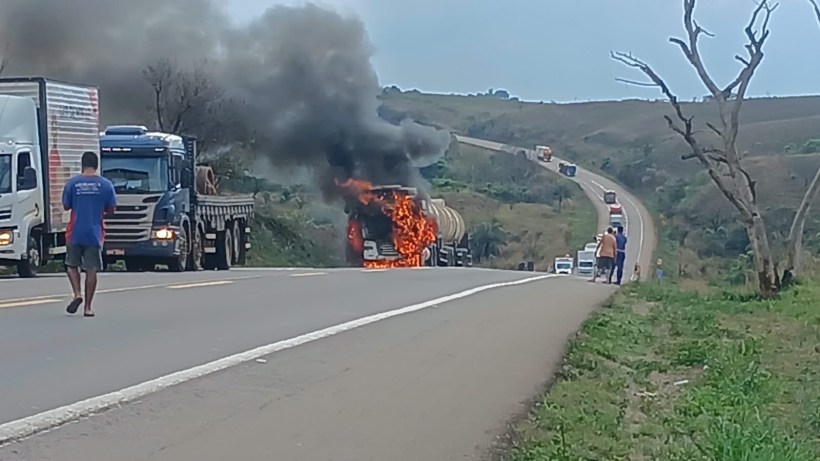 Caminhão pegando fogo em uma estrada, com fumaça e chamas visíveis. Outras caminhões e pessoas estão tentando se afastar da cena.