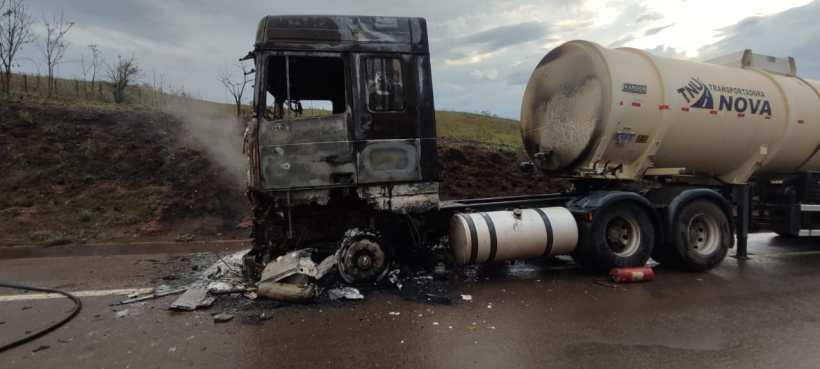 Caminhão danificado por incêndio, com a cabine queimada e fumaça saindo. O tanque de transporte está ao lado na estrada.