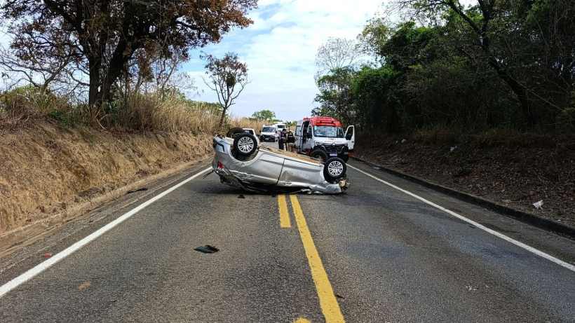 Um carro capotado na estrada, com equipes de emergência ao fundo, em um cenário rural.