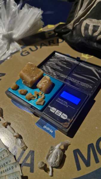 Uma balança digital com pedaços de substância sobre ela, em cima de uma superfície que diz 'Guarda'. Ao fundo, sacos plásticos e uma mochila.