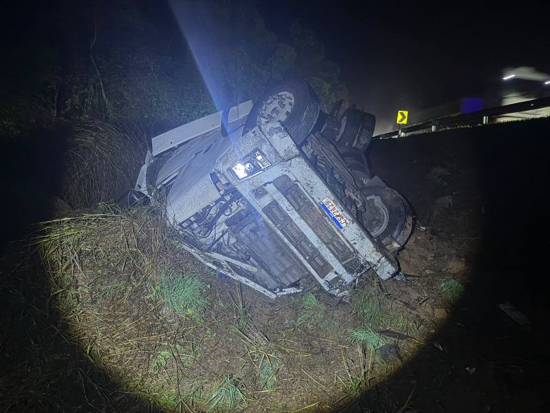 Caminhão tombado em um terreno ao lado de uma estrada à noite, com faróis iluminando a área.