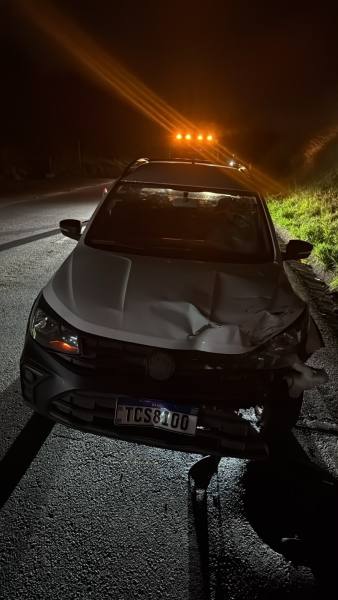 Carro acidentado na estrada à noite, com a parte da frente danificada e luzes de sinalização ao fundo.