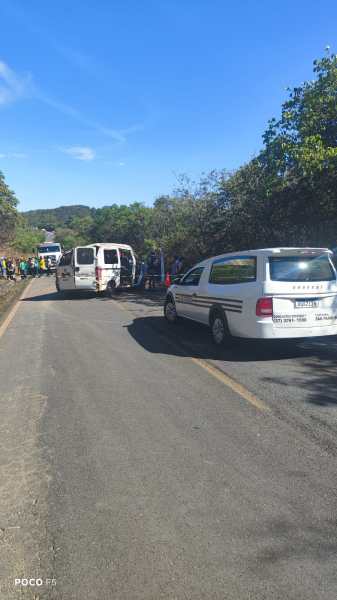 Cena de uma estrada com veículos parados, incluindo uma van branca e um carro, cercados por vegetação. Há um grupo de pessoas ao fundo, sugerindo uma atividade ou evento.
